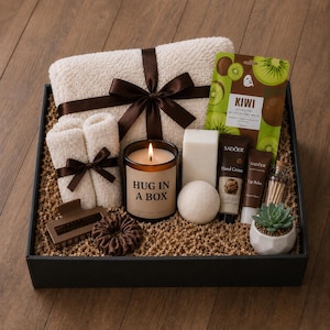 May include: A self-care gift box containing a white towel with a brown ribbon, a lit candle with the text "HUG IN A BOX", and beauty products. The box also includes a small succulent in a white pot, offering a spa-like experience.