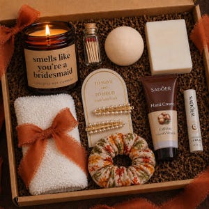 May include: A gift box containing a candle, matches, soap, hair accessories, hand cream, lip balm, and a washcloth. The candle reads "smells like you're a bridesmaid." The box has a fall theme with orange and brown accents.