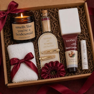 May include: A gift box containing a candle, matches, hair accessories, a towel, hand cream, and lip balm. The candle reads "smells like you're a bridesmaid." The box is decorated with a burgundy ribbon and rose petals.
