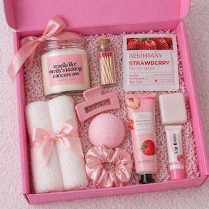 May include: A pink gift box containing self-care products. It features a candle with text, matches, a hair clip, a bath bomb, a scrunchie, a facial mask, hand cream, soap, and lip balm. The box is tied with a pink ribbon.