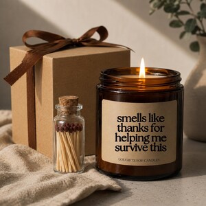 May include: A lit soy candle in a brown glass jar with the text "smells like thanks for helping me survive this." A small glass bottle of matches and a brown gift box with a ribbon are also present.