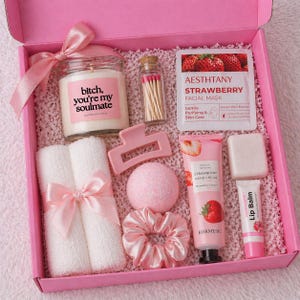 Peut inclure: Coffret cadeau rose rempli d'articles de bien-être. Il comprend une bougie avec du texte, un masque facial, une crème pour les mains, un baume à lèvres, une pince à cheveux, une bombe de bain, un chouchou, des allumettes et deux petites serviettes blanches avec un nœud rose.