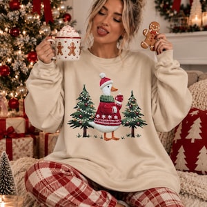 Peut inclure: Un sweat-shirt beige à col rond orné d'un motif représentant un canard blanc vêtu d'un pull en tricot rouge et vert avec un bonnet assorti, debout entre deux sapins de Noël décorés. Le canard tient une petite tasse rouge dans son aile.
