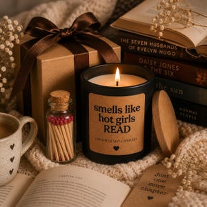 Peut inclure: Une bougie en cire de soja noire dans un pot en verre avec une étiquette beige portant l'inscription "smells like hot girls READ" et un petit cœur. La bougie est allumée à côté d'un flacon en verre de allumettes à bouts rouges et de livres.