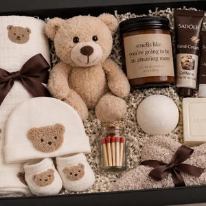 May include: A gift box containing baby essentials and self-care items. It features a teddy bear, blanket, hat, and booties, all with bear motifs. Also includes a candle, hand cream, lip balm, bath bomb, soap, and matches. The candle reads "smells like you're going to be an amazing mom."