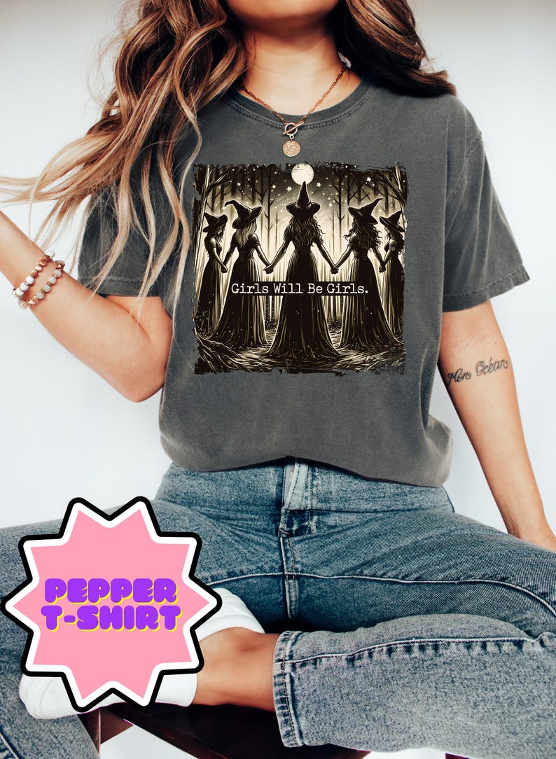 May include: Dark gray t-shirt featuring a graphic of five witches holding hands in a forest, with the text "Girls Will Be Girls." The shirt is part of the Pepper T-Shirt collection.