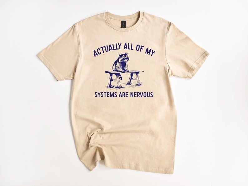 Puede incluir: Camiseta color beige con el texto azul marino &laquo;ACTUALLY ALL OF MY SYSTEMS ARE NERVOUS&raquo;. Una ilustraci&oacute;n de mapache est&aacute; sentada en un banco. La camiseta est&aacute; colocada sobre un fondo blanco.