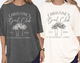 May include: Two oversized t-shirts, one charcoal and one ivory, with the text "CHRISTINE'S Bach Club" and a palm tree graphic. The text "EST. 2026" and "ST. PETERSBURG, FLORIDA" are below the graphic.