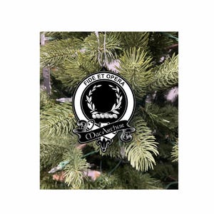 May include: A black and white Macarthur family crest Christmas tree ornament. The ornament features the words "Fide et Opera" and "MacArthur". The round ornament has a wreath design and is suspended from a tree.
