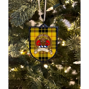 Peut inclure: Une décoration de Noël en tartan jaune et noir en forme de bouclier. Le bouclier présente une crête rouge et bleue avec une tête de taureau et le texte "Hold Fast" et "MacLeod".