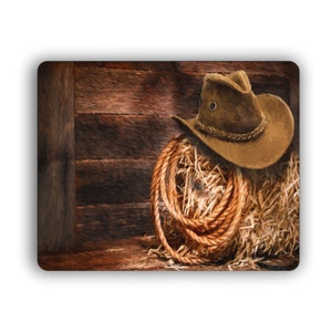 May include: A brown cowboy hat with a brown leather band sits on a pile of hay with a brown rope wrapped around it. The hay and rope are against a wooden wall.