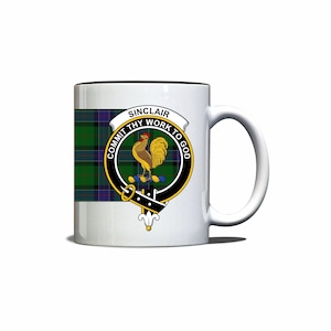 May include: White ceramic mug with a black rim. The mug features a green, blue, and black tartan pattern with a crest in the center. The crest depicts a rooster with the text "Sinclair Commit Thy Work To God".
