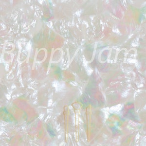 Op de afbeelding: Een wit iriserend materiaal met een gekreukelde, reflecterende textuur die subtiele regenboogaccenten vertoont. De tekst Buppy Jam Ltd is in het wit over het midden geplaatst. Drie kleine, dunne, lichtgekleurde verticale objecten zijn zichtbaar.