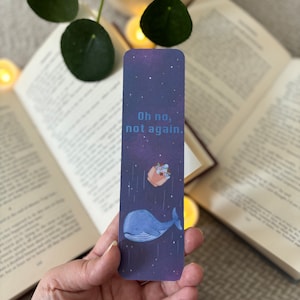Peut inclure: Un marque-page violet avec une baleine bleue et un petit pot de fleurs tombant du ciel. Le texte sur le marque-page est "Oh non, pas encore."