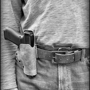 以下が含まれることがあります： 革製のホルスターに入った黒い拳銃を、人がベルトに付けています。ベルトは茶色の革製で、銀色のバックルが付いています。その人は青いジーンズと灰色のシャツを着ています。