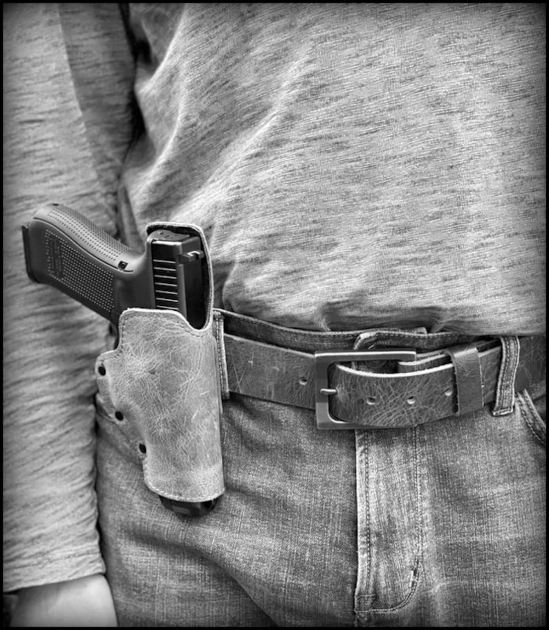 以下が含まれることがあります： 革製のホルスターに入った黒い拳銃を、人がベルトに付けています。ベルトは茶色の革製で、銀色のバックルが付いています。その人は青いジーンズと灰色のシャツを着ています。
