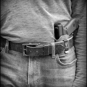 以下が含まれることがあります： 黒い革のベルトに銀色のバックルと黒い銃ホルスターが取り付けられています。ホルスターには黒い銃が入っており、小さな黒いラベルに「VF」という文字が書かれています。