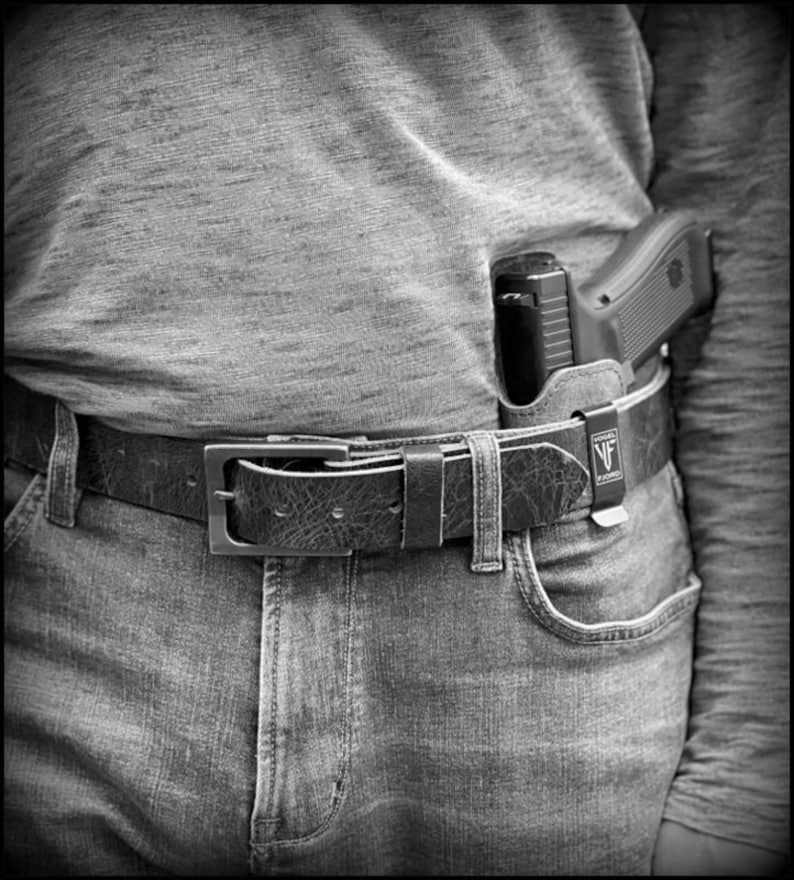以下が含まれることがあります： 黒い革のベルトに銀色のバックルと黒い銃ホルスターが取り付けられています。ホルスターには黒い銃が入っており、小さな黒いラベルに「VF」という文字が書かれています。