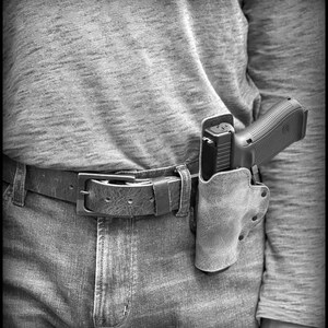 以下が含まれることがあります： 革製のホルスターに収められた黒い拳銃。茶色の革製のベルトで腰に装着されている。