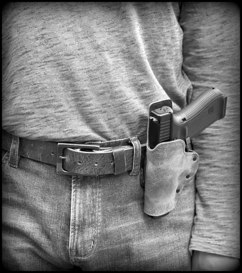 以下が含まれることがあります： 革製のホルスターに収められた黒い拳銃。茶色の革製のベルトで腰に装着されている。