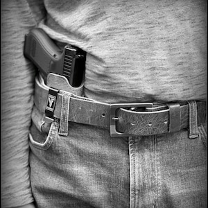 以下が含まれることがあります： 黒い銃がホルスターに収められ、銀色のバックルが付いた茶色の革のベルトに固定されています。ベルトは青いジーンズの上に着用されています。