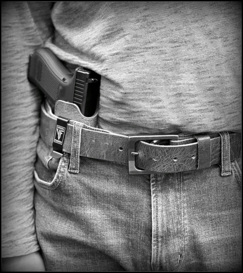 以下が含まれることがあります： 黒い銃がホルスターに収められ、銀色のバックルが付いた茶色の革のベルトに固定されています。ベルトは青いジーンズの上に着用されています。