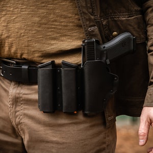 May include: A black leather gun holster with a magazine carrier attached to a brown leather belt. The holster is designed to hold a handgun and extra ammunition.