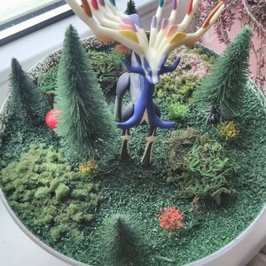 May include: A miniature forest scene with a colorful, multi-colored, deer-like creature with antlers in the center. The creature is surrounded by small, green, artificial trees and moss.