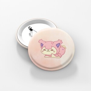 May include: Two round, light pink buttons. One button features a cartoon cat in a pink hooded outfit with purple accents. The other button is plain with a silver pin back. The buttons are on a white surface.