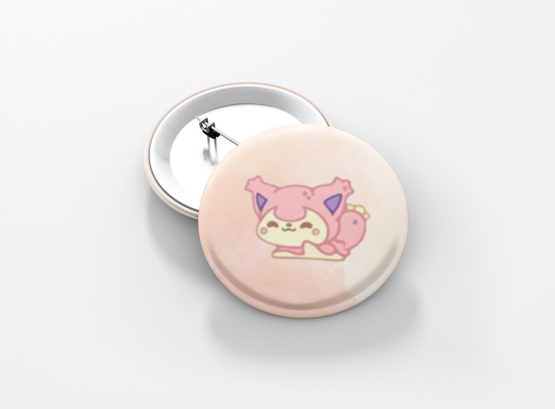 May include: Two round, light pink buttons. One button features a cartoon cat in a pink hooded outfit with purple accents. The other button is plain with a silver pin back. The buttons are on a white surface.