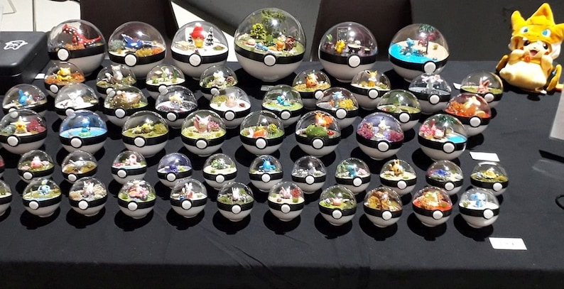 May include: A collection of 35 clear glass Poke Balls, each containing a miniature diorama with various Pokemon characters and greenery. The Poke Balls are arranged on a black surface.