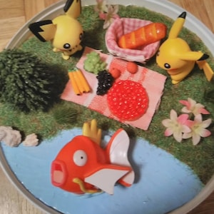 May include: A miniature scene of a picnic with two yellow Pokemon characters, a red fish Pokemon, a small green tree, and a picnic basket with a loaf of bread. The scene is set on a round, white base with a blue pond and green grass.