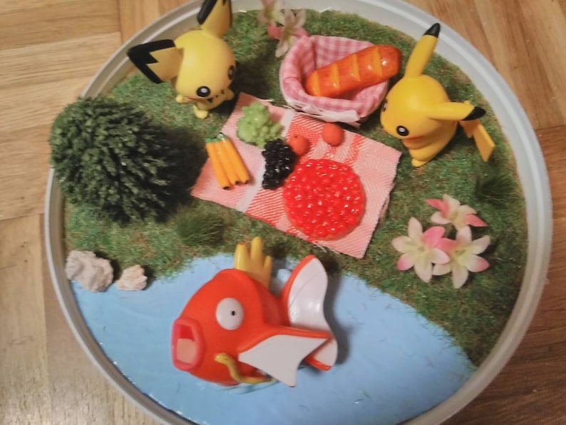 May include: A miniature scene of a picnic with two yellow Pokemon characters, a red fish Pokemon, a small green tree, and a picnic basket with a loaf of bread. The scene is set on a round, white base with a blue pond and green grass.