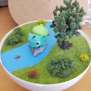 May include: A miniature diorama featuring a green Bulbasaur Pokemon figurine on a rock in a blue river, surrounded by green grass, a small tree, and other miniature plants.