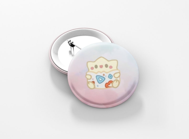 May include: Two round, light pink pinback buttons. One button is lying flat, and the other is upright. The buttons feature a cartoon Togepi character on a pastel pink and blue background.