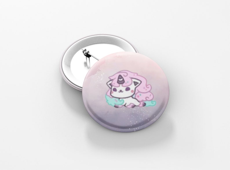 May include: Two round, white-backed buttons. One button features a cartoon unicorn-cat with pink hair, a purple horn, and a light blue cloud. The background is a gradient of pink and purple with white speckles. The second button is blank.
