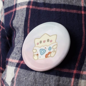 May include: A round, glossy button with a cartoon Togepi design. The button features a light pink and blue gradient background. The Togepi character is in the center, with a white, blue, and orange color scheme. The button is set against a plaid shirt.