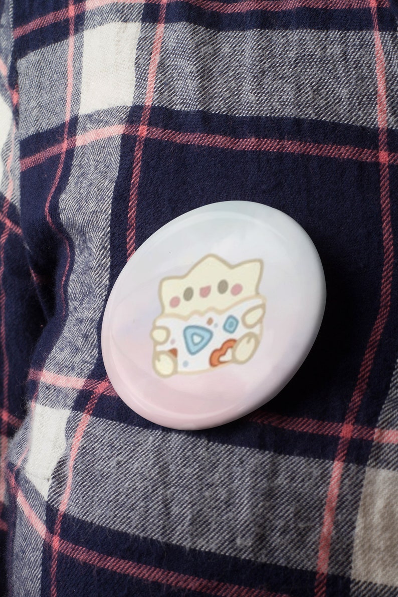 May include: A round, glossy button with a cartoon Togepi design. The button features a light pink and blue gradient background. The Togepi character is in the center, with a white, blue, and orange color scheme. The button is set against a plaid shirt.