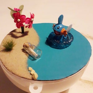 May include: A miniature beach scene with a blue and orange Pokemon figurine in the water, a message in a bottle on the sand, and a pink flower arrangement.