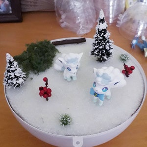 May include: A white and blue Pokemon figurine with a fluffy white tail and a blue bow. The figurine is standing on a white surface with a small green bush and a miniature snow-covered pine tree.