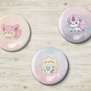 May include: Three round, colorful buttons with cartoon illustrations. One button features a pink creature, another a purple unicorn-like character, and the third a yellow and white creature. The buttons are set against a light wood grain background.