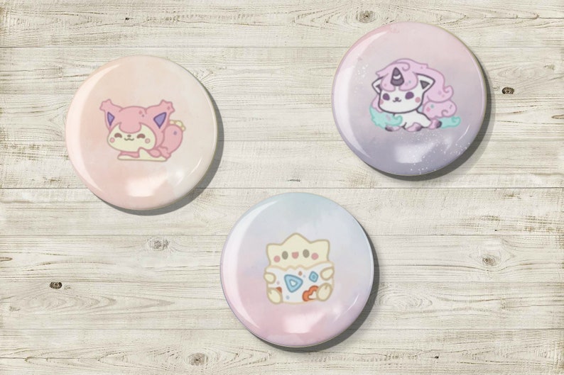 May include: Three round, colorful buttons with cartoon illustrations. One button features a pink creature, another a purple unicorn-like character, and the third a yellow and white creature. The buttons are set against a light wood grain background.