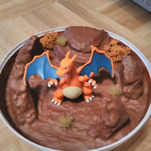 May include: A brown and orange Pokemon figurine with blue wings standing on a brown, rocky, and textured surface. The surface is in a white bowl.