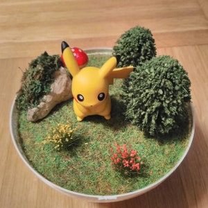 May include: A yellow Pikachu figurine standing in a miniature garden scene. The scene includes a small rock, a red mushroom, and two green bushes. The garden is surrounded by green grass.