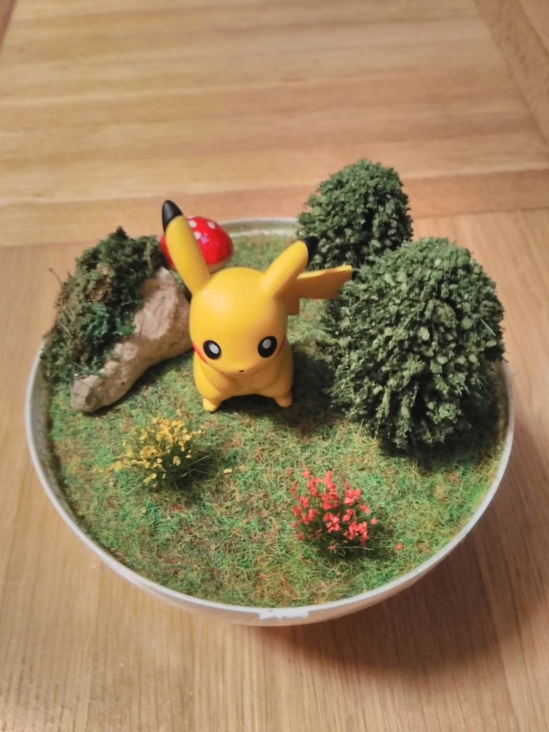 May include: A yellow Pikachu figurine standing in a miniature garden scene. The scene includes a small rock, a red mushroom, and two green bushes. The garden is surrounded by green grass.