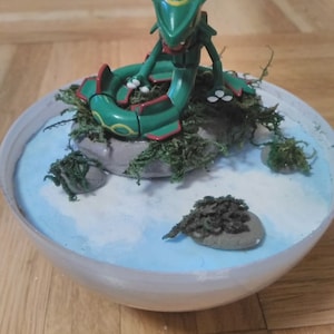 May include: A green and yellow Pokemon figure of a serpentine creature sitting on a small rock island in a white and blue bowl. The bowl is filled with white and blue paint and has a small amount of green moss around the edges.