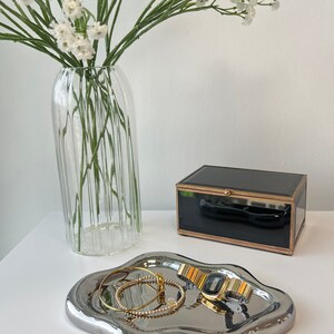 May include: A silver, wavy, decorative tray with a gold watch and two gold bracelets on top. The tray is sitting on a white surface next to a clear glass vase with white flowers and a black glass box with a gold trim.