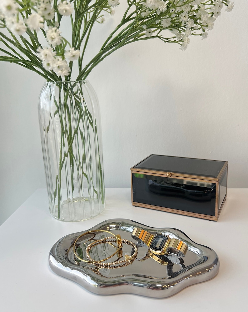 May include: A silver, wavy, decorative tray with a gold watch and two gold bracelets on top. The tray is sitting on a white surface next to a clear glass vase with white flowers and a black glass box with a gold trim.
