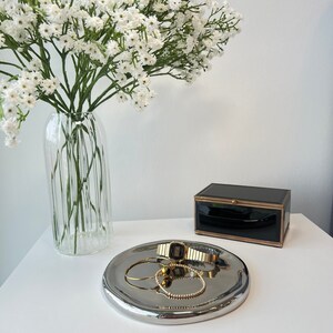 May include: A silver tray with a gold watch, a gold bracelet, and a gold ring on a white surface. A vase with white flowers is in the background.