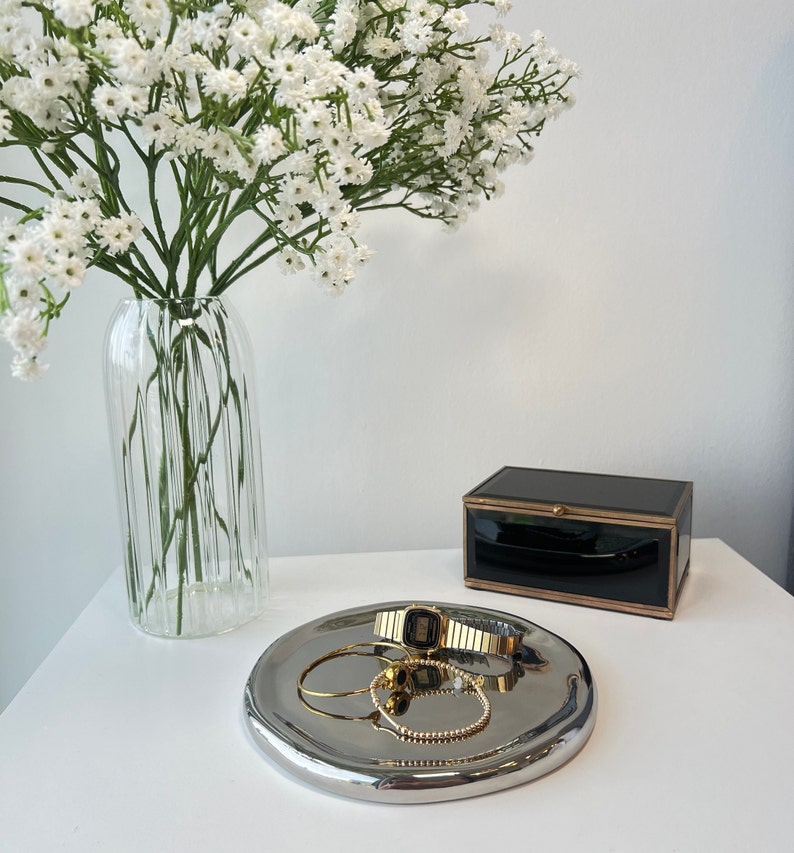 May include: A silver tray with a gold watch, a gold bracelet, and a gold ring on a white surface. A vase with white flowers is in the background.